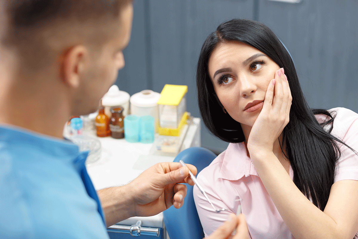 3-Signs-You-Need-a-Root-Canal woman patient holds her hand to the side of her jaw while in pain while the dentist tells her she needs a root canal