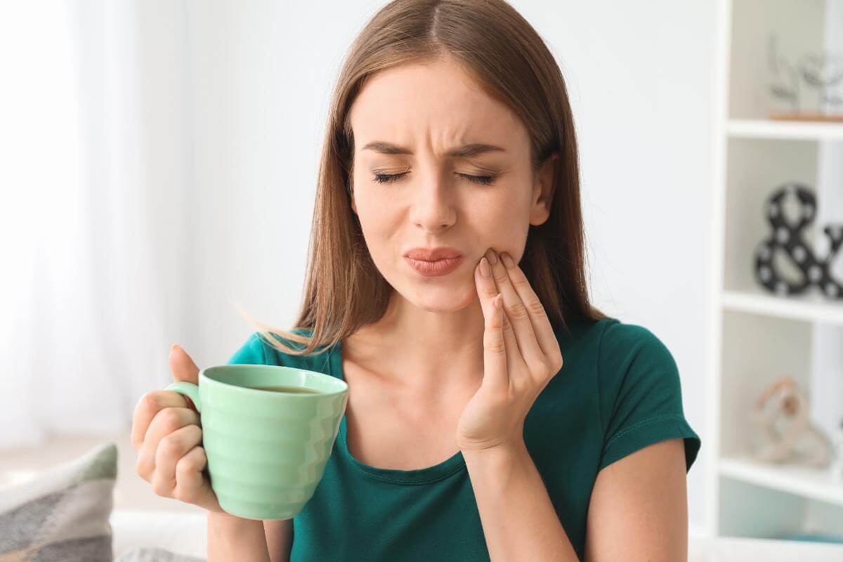 a woman holds onto her coffee mug with one hand and the other hand holds her jaw in pain because of a dental cavity