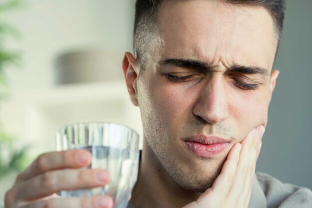 a man holds a glass of water while his other hand holds onto the side of his mouth in pain and realizing he needs to improve his gum health