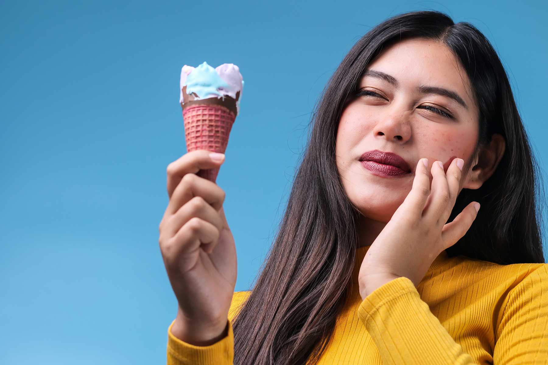 a woman holds an ice cream cone in one hand and raises her other hand to her jaw while experiencing pain in her sensitive teeth