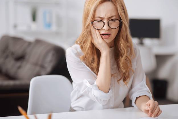 What-Are-the-Signs-of-a-Tooth-Infection woman sitting at a desk in pain holds onto the side of mouth as she experiences signs of a tooth infection