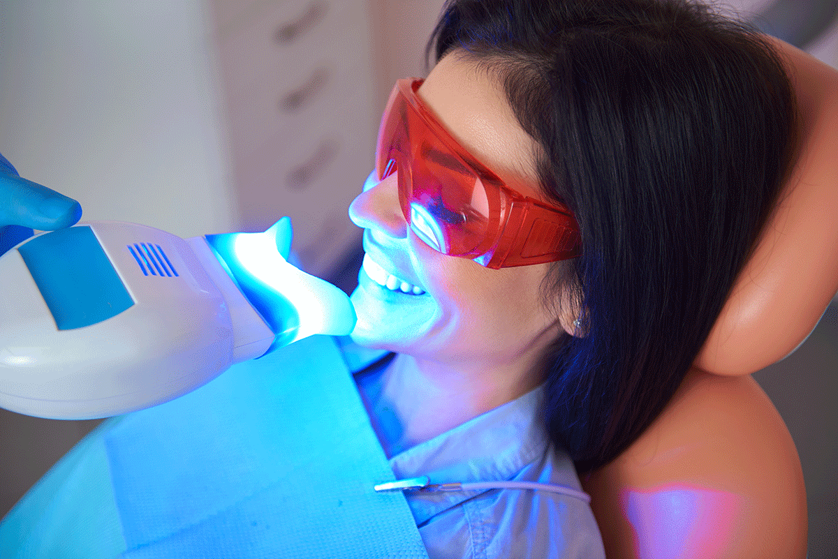 a woman patient sits in a dental exam chair with teeth whitening light placed near her mouth while she asks about the cost of teeth whitening