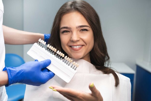 a dental professional holds up dental implant shades while patient sits in exam chair and smiles and asks dentist about how to care for veneers