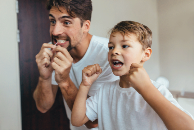What-Are-the-Benefits-of-Flossing-Your-Teeth a dad and his son stand in front of a mirror flossing their teeth together while the dad tells the son about the benefits of flossing