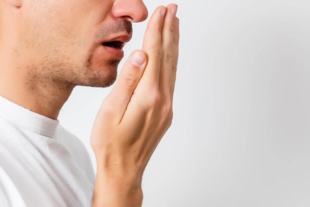 Common Causes of Bad Breath man holds his hand to his mouth and breathes while wondering about the common causes of bad breath