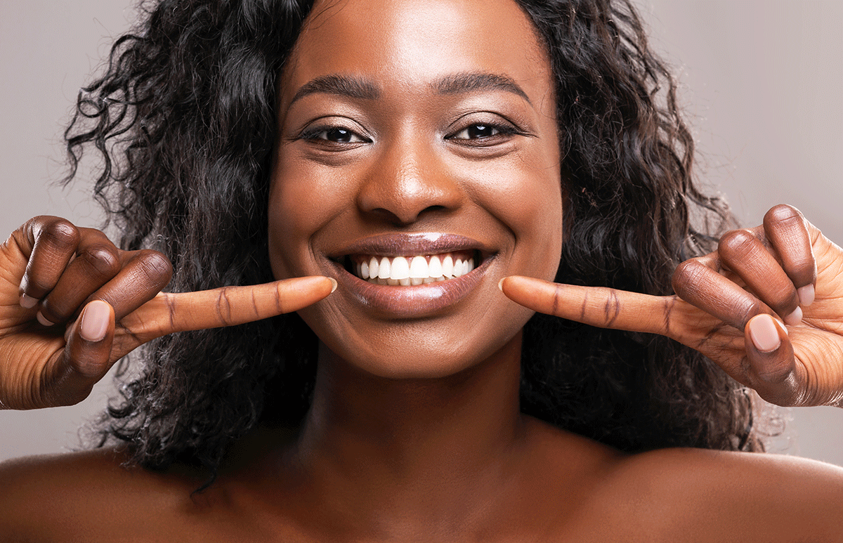 What-Do-Healthy-Gums-Look-Like woman smiles and points both of her index fingers to her smile after answering the question of what do healthy gums look like