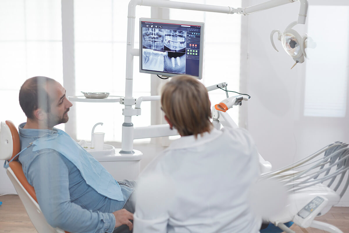 a dentist sits with a patient in an exam room explaining to him the pro and cons of dental implants