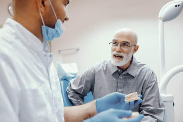 5 Benefits Of Choosing Dental Implants Over Dentures older man with a white beard and glasses sits in dental exam room and asks about the benefits of dental implants to dental expert