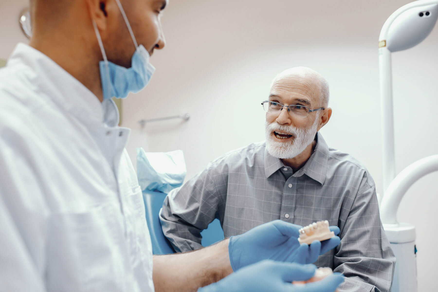 5 Benefits Of Choosing Dental Implants Over Dentures older man with a white beard and glasses sits in dental exam room and asks about the benefits of dental implants to dental expert