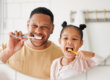 a dad and his daughter brush their teeth after learning how to prevent cavities while looking at their reflections in a mirror