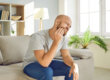 man sitting on a couch holds his hand to the side of his face near his jaw showing signs of a dental emergency