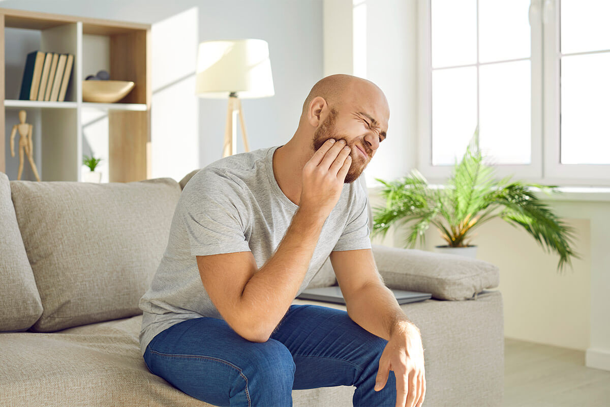 man sitting on a couch holds his hand to the side of his face near his jaw showing signs of a dental emergency