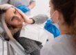 A woman sitting in a dental chair holds her cheek in pain while speaking with a dentist wearing gloves and a mask.