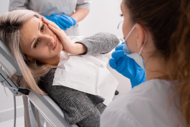 beautiful caucasian girl with toothache sits in a dental chair. Professional dentist helps his female patient in pain.painful treatment help. A woman sitting in a dental chair holds her cheek in pain while speaking with a dentist wearing gloves and a mask.