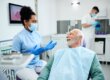 A female dentist wearing blue scrubs and a mask talks to an older male patient in a dental chair while another staff member works in the background.