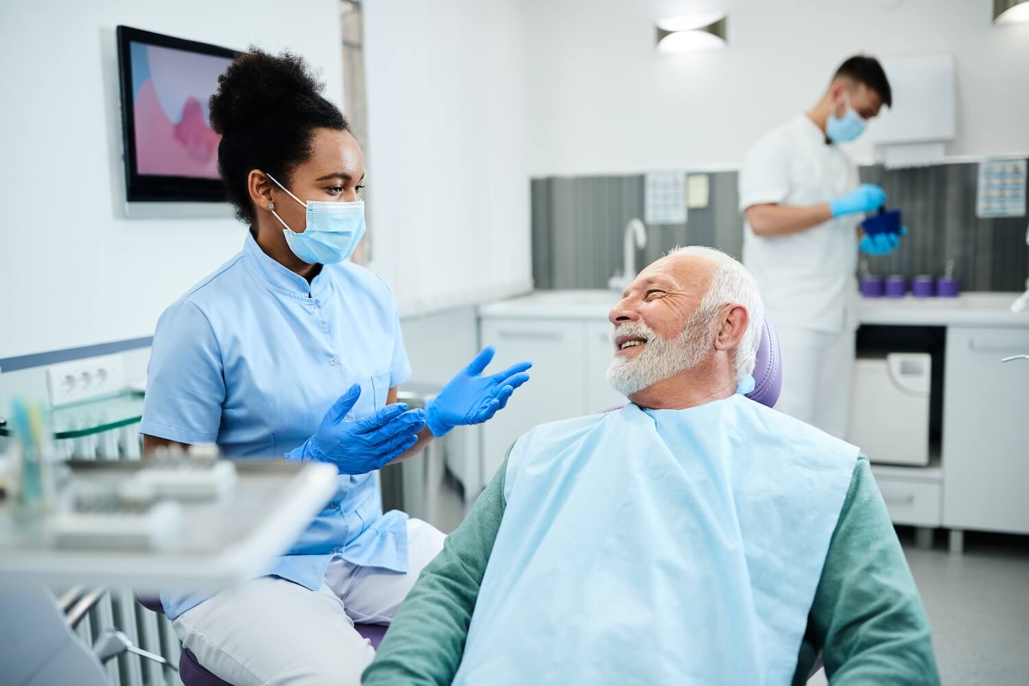 A female dentist wearing blue scrubs and a mask talks to an older male patient in a dental chair while another staff member works in the background.