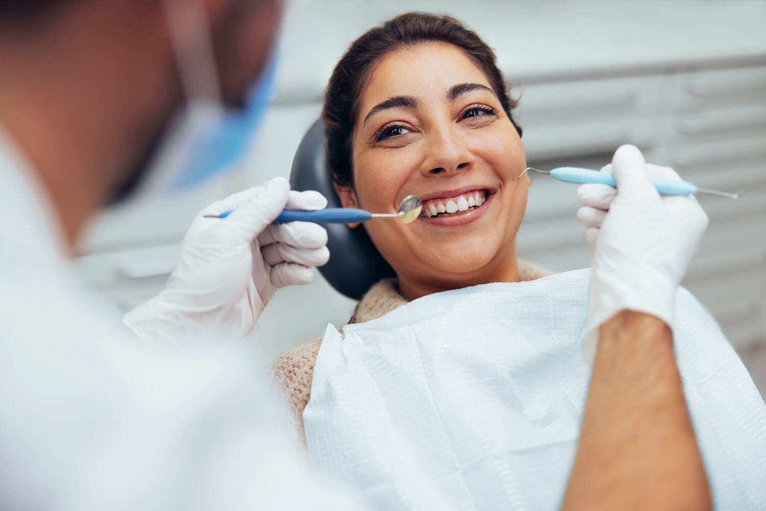 A smiling woman sitting in a dental chair during a check-up as a dentist holds dental tools near her mouth.