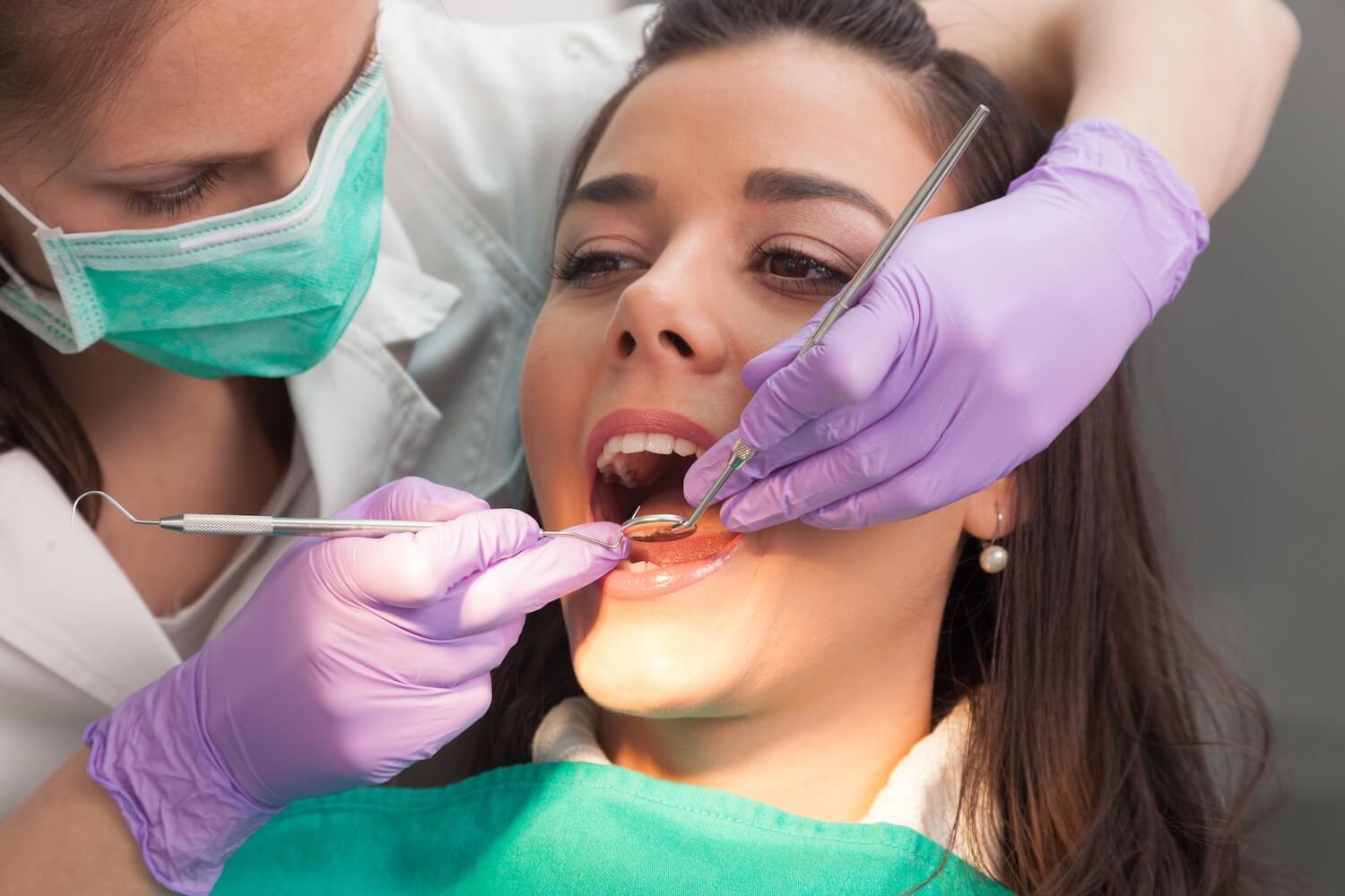 Best Ways to Keep Your Natural Teeth From Fluoride Treatments to Root Canal Therapy A dentist wearing a mask and purple gloves examines a woman’s open mouth using dental tools during a check-up.