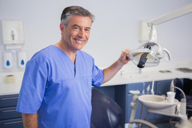 Finding a Local Dentist Near You in Houston at Supreme Dentistry A smiling dentist in blue scrubs stands beside a dental chair, adjusting an overhead examination light in a clean, modern dental office.