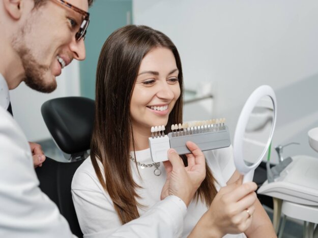 A woman smiles while holding a mirror as a dentist compares tooth shade samples next to her teeth during a cosmetic consultation.