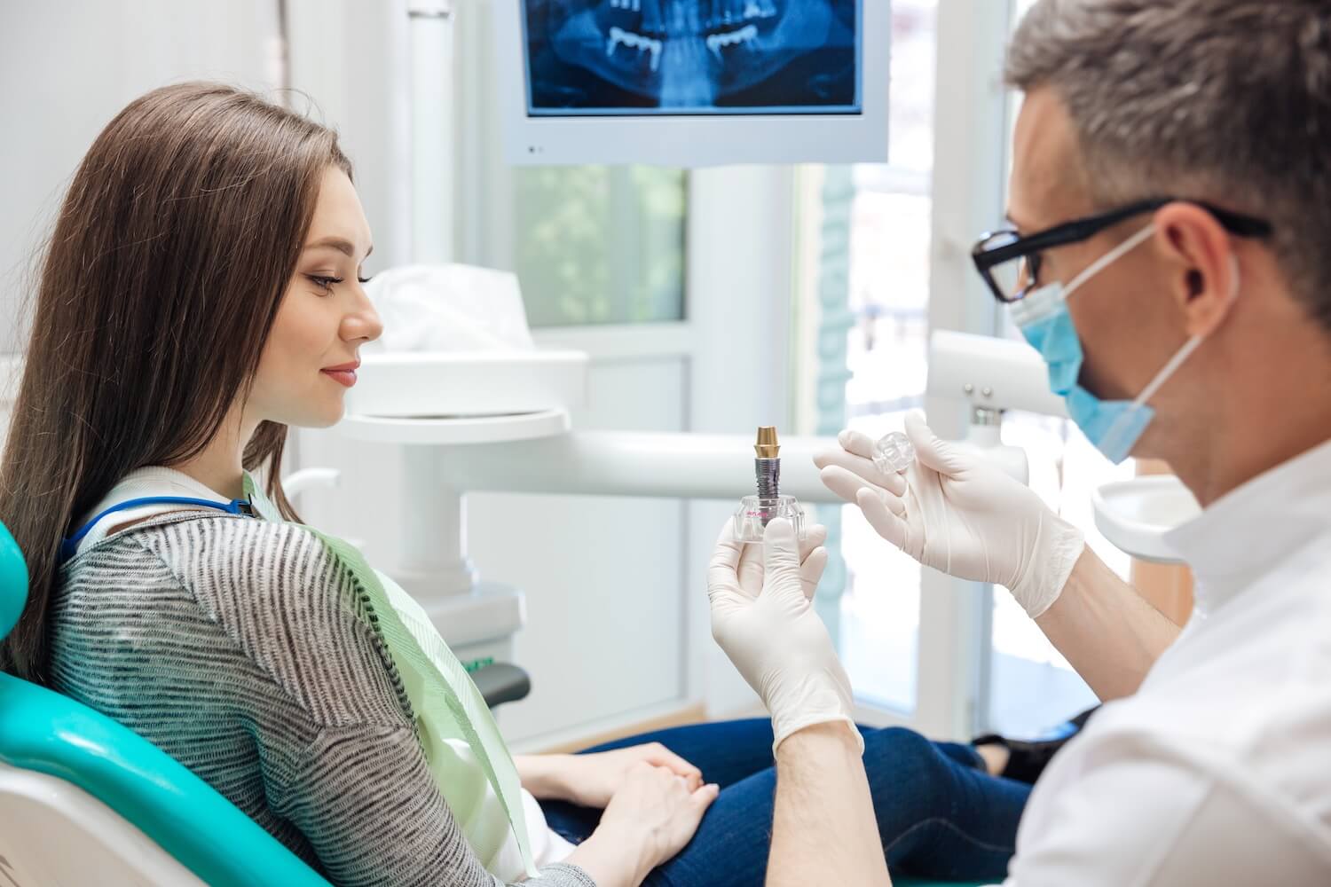 Male dentist showing his female patient a dental implant in clinic A woman sits in a dental chair while a dentist wearing gloves and a mask shows her a model of a dental implant, with a dental X-ray displayed on a monitor in the background.