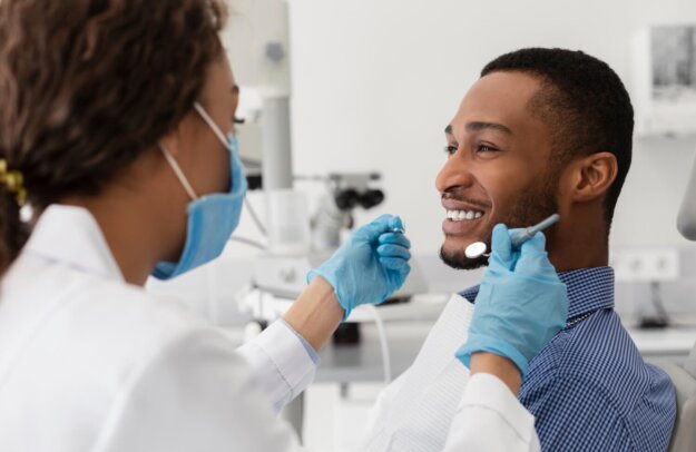 Why Fluoride Treatment Is a Must in Cypress A man sits in a dental chair smiling while a dentist wearing gloves and a face mask holds dental instruments and prepares to examine his teeth.